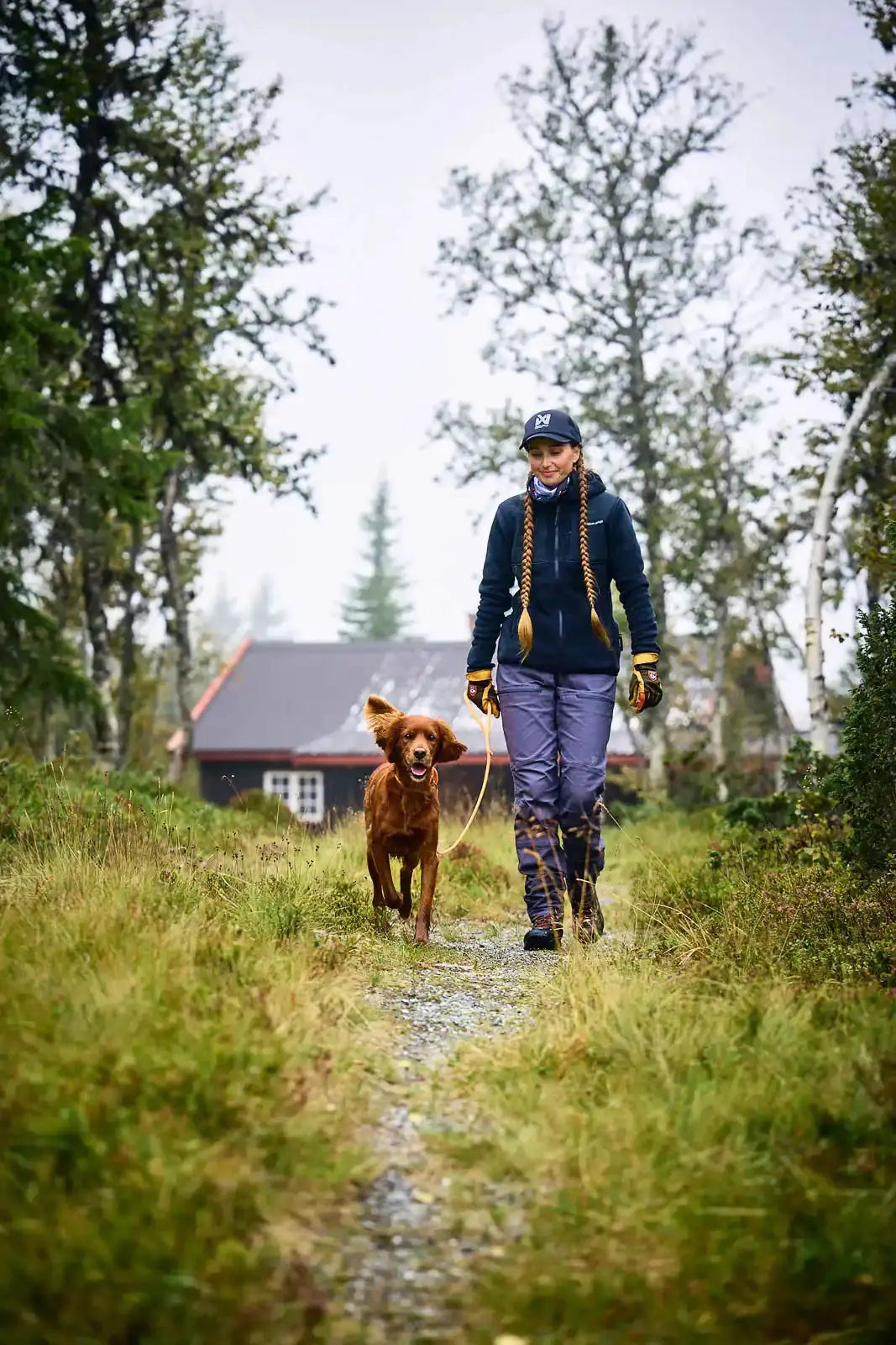 Person walking a dog on a path with trees and a building in the background