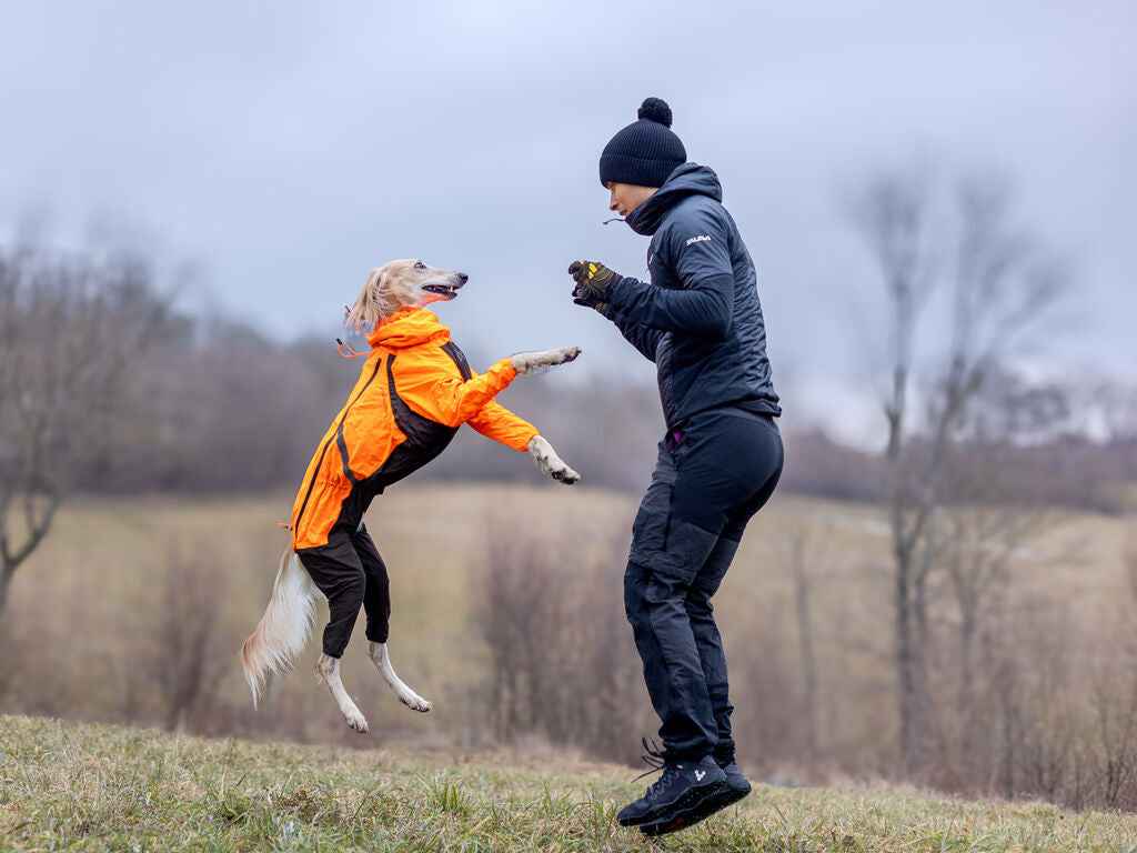A person in a black jacket and hat is jumping up to catch a dog wearing an orange jacket, both in a grassy field.