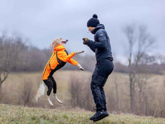 A person in a black jacket and hat is jumping up to catch a dog wearing an orange jacket, both in a grassy field.