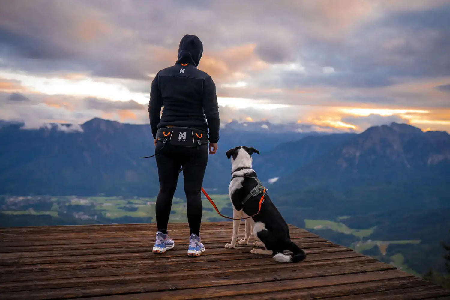 Person and dog standing on a wooden platform overlooking mountains with a sunset or sunrise.